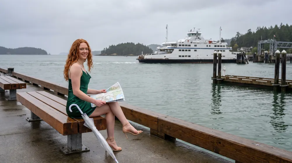 Isla Whitcombe sits on a waterfront bench with a road map while a car ferry crosses the harbour behind her. A character created for the guide; AI-generated image.